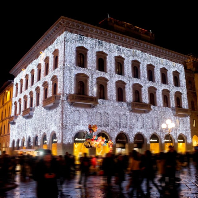Façade d’un bâtiment couverte de rideaux lumineux blancs, avec passants sur la place de nuit
